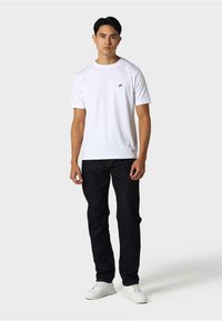 Bela kratka majica s kratkimi rokavi in krožnim izrezom ter majhnim logotipom na prsih, v kombinaciji s temnimi kavbojkami in belimi teniskami.