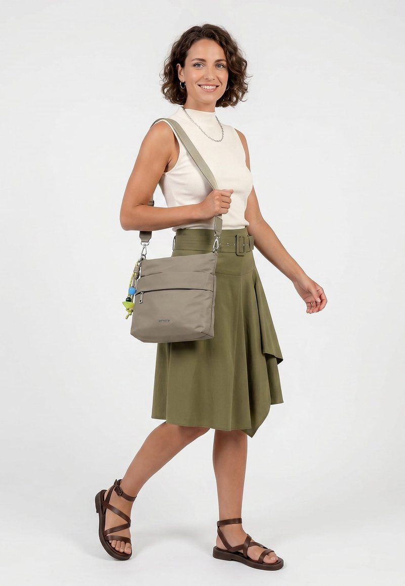 Lachende vrouw die een mouwloze witte top draagt, olijfgroene rok met riem, bruine sandalen, draagt een beige schoudertas met decoratieve sleutelhanger.