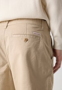 Béžové bavlněné kalhoty s zadní kapsou se zapínáním na knoflík a červenobílou etiketou Calvin Klein. Hladká textura a přizpůsobený střih.