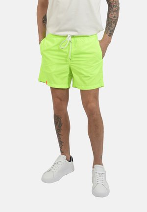 Uomo con pantaloncini verde neon con coulisse, sneakers bianche, t-shirt bianca, con tatuaggi visibili su braccia e gambe, in piedi con le mani nelle tasche.