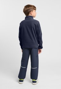 Pullover in pile navy con colletto alto abbinato a pantaloni navy con strisce riflettenti e sneakers con dettagli verdi.