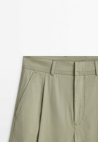 Pantalons plissés vert clair avec une taille plate, des poches latérales et des détails de couture contrastés, fabriqués en tissu lisse et léger.