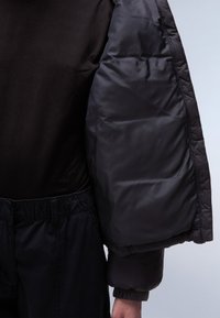 Dikke zwarte jas met brede doorgestikte mouwen, gecombineerd met een zwarte top en zwarte broek met elastische taille. Zachte textuur en minimalistisch ontwerp.