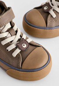 Braune High-Top-Sneaker mit runder Zehenpartie, beigen Schnürsenkeln und einem bärenförmigen Akzent an der Oberseite. Texturierte Gummisohle mit griffiger Oberfläche.