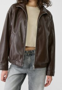 Chaqueta bomber de cuero marrón con cremallera frontal, diseño recortado, bolsillos laterales y dobladillo acanalado. Se lleva sobre un top corto beige y vaqueros.