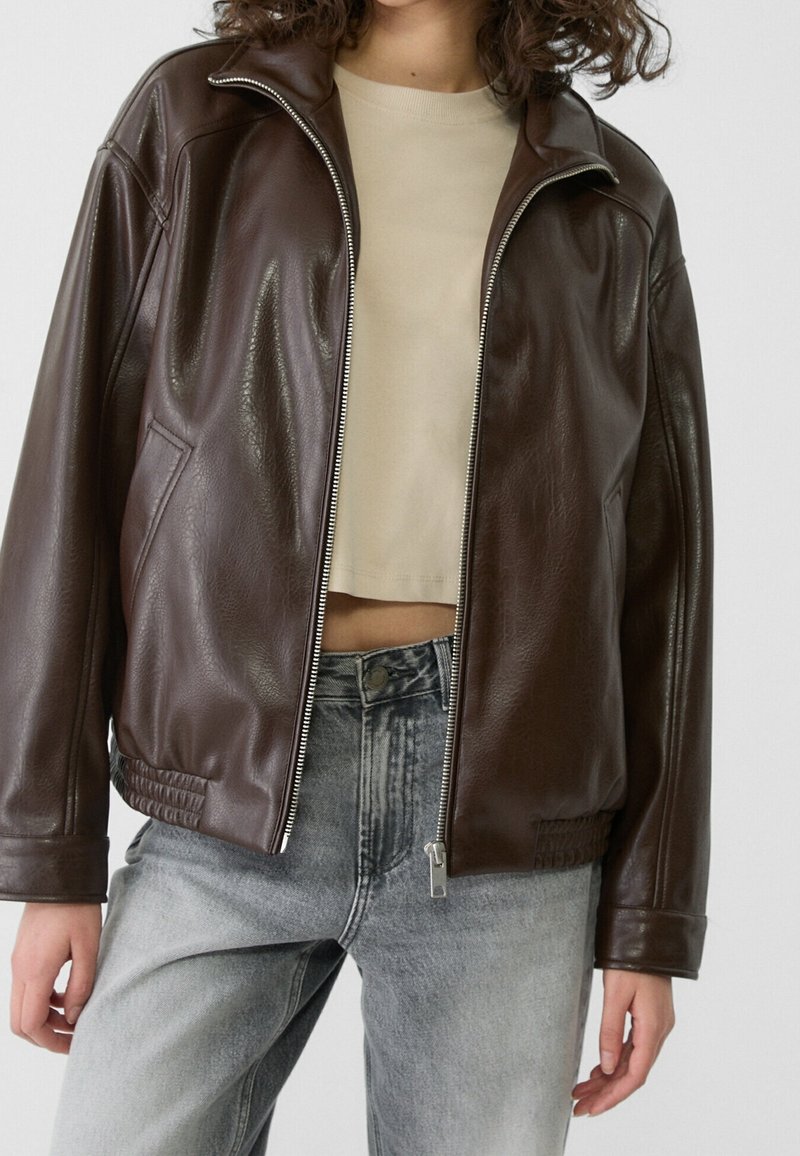Chaqueta bomber de cuero marrón con cremallera frontal, diseño recortado, bolsillos laterales y dobladillo acanalado. Se lleva sobre un top corto beige y vaqueros.