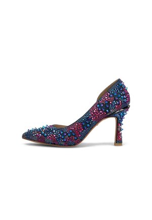 Zapato de tacón alto puntiagudo cubierto de picos y tachuelas iridiscentes azules y rosas sobre un fondo blanco.