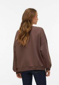 Sudadera marrón de gran tamaño con hombros caídos, puños y bajo acanalados. Se lleva con vaqueros azul oscuro, con una textura suave.