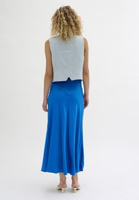Eine Frau mit lockigem blonden Haar trägt ein ärmelloses hellblaues Oberteil, einen leuchtend blauen Midiskirt und weiße High Heels und steht vor einem schlichten weißen Hintergrund.