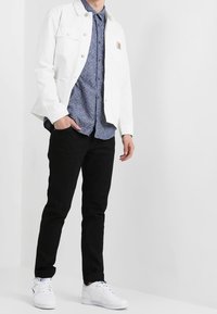 Witte spijkerjacket over een blauw patroonshirt, gecombineerd met zwarte broek en witte sneakers. Kenmerken zijn onder andere twee voorzakken en een logopatch.