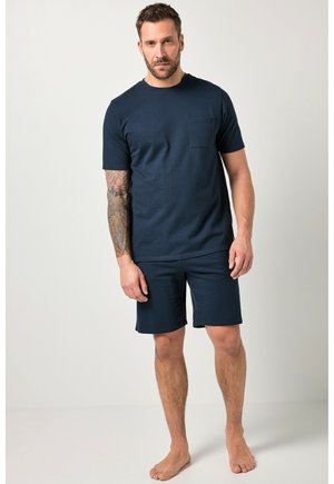 Uomo con braccio tatuato che indossa una maglietta blu navy a maniche corte e pantaloncini abbinati, in piedi a piedi nudi contro uno sfondo chiaro e uniforme.