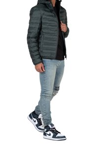 Uomo con giacca trapuntata verde scuro, camicia nera, jeans chiari strappati con testo e sneakers alte bianco e nere, in piedi di lato.