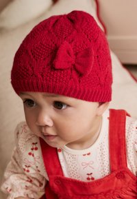 Bonnet tricoté rouge avec un motif texturé et un nœud décoratif. Conçu pour les nourrissons, il offre un ajustement serré et un matériau doux pour le confort.