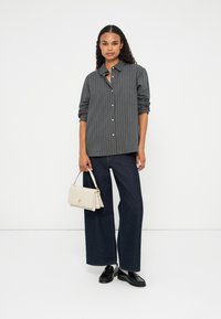 Camicia grigia a righe con bottoni, pantaloni larghi in denim scuro, mocassini neri e una borsa color crema con un accento dorato.