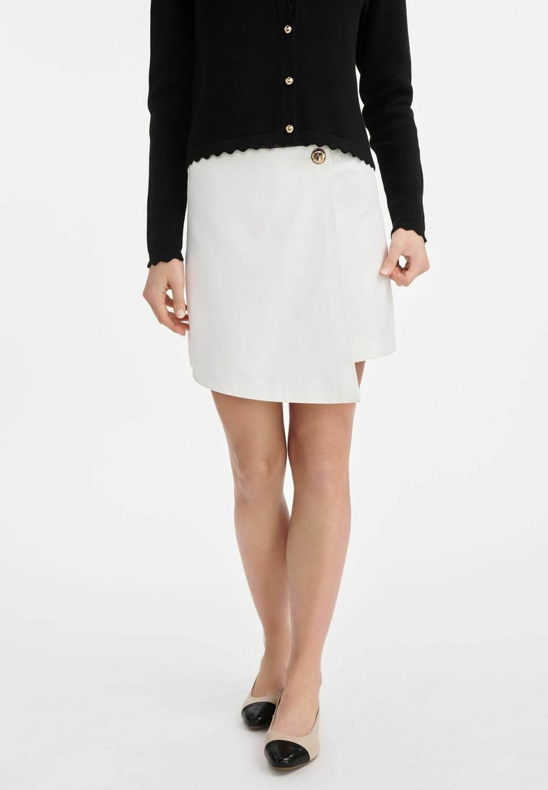 Biała spódnica o kroju owijanym z bocznym zapięciem na guziki, noszona z czarnym swetrem z długim rękawem. Spódnica ma gładką fakturę i lekki połysk.