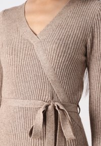 Robe en tricot côtelé beige avec décolleté en V, manches longues et taille nouée. Texture douce avec des détails en côtes verticales sur l'ensemble.
