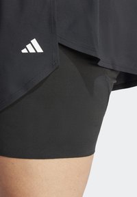 Černé sportovní šortky z hladké, pružné látky s vrstveným černým topem a bílým trojúhelníkovým logem jako akcentem.