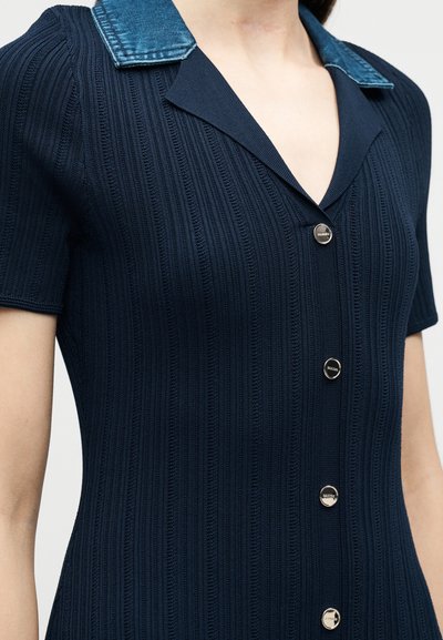 Φόρεμα από πλεκτό ύφασμα σε ναυτικό μπλε με ριμπ υφή, κοντά μανίκια, τέσσερα μεταλλικά κουμπιά στο μπροστινό μέρος και αντίθετο γιακά από μπλε τζιν.