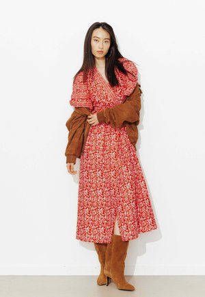 Femme portant une robe midi à motifs rouges et blancs, une veste en daim marron drapée sur les épaules, et des bottes en daim marron assorties montantes jusqu'aux genoux.