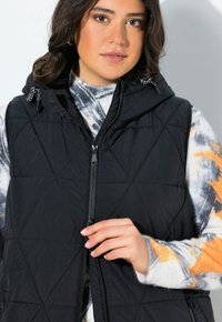 Svart quiltad väst med huva, försedd med dragkedja framtill och snörning. Bärs över en mönstrad tröja i orange, grå och vita toner.