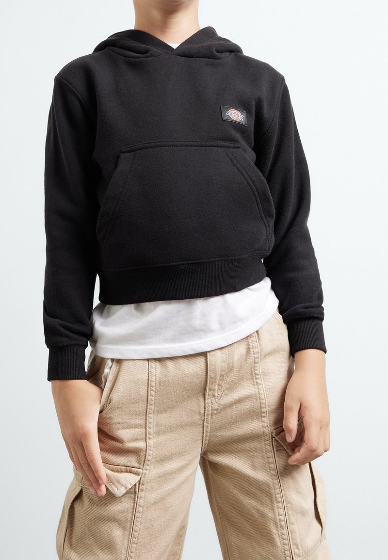 Enfant portant un sweat à capuche noir avec une poche avant et un pantalon cargo beige, debout devant un fond clair uni.