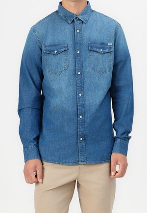 Homme portant une chemise en jean bleue à boutons avec deux poches poitrine et un pantalon beige, debout les mains relâchées le long du corps.