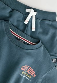 Felpa outdoor Superdry blu-verde e pantaloni abbinati con cordoncino bianco, piegati su una superficie chiara.