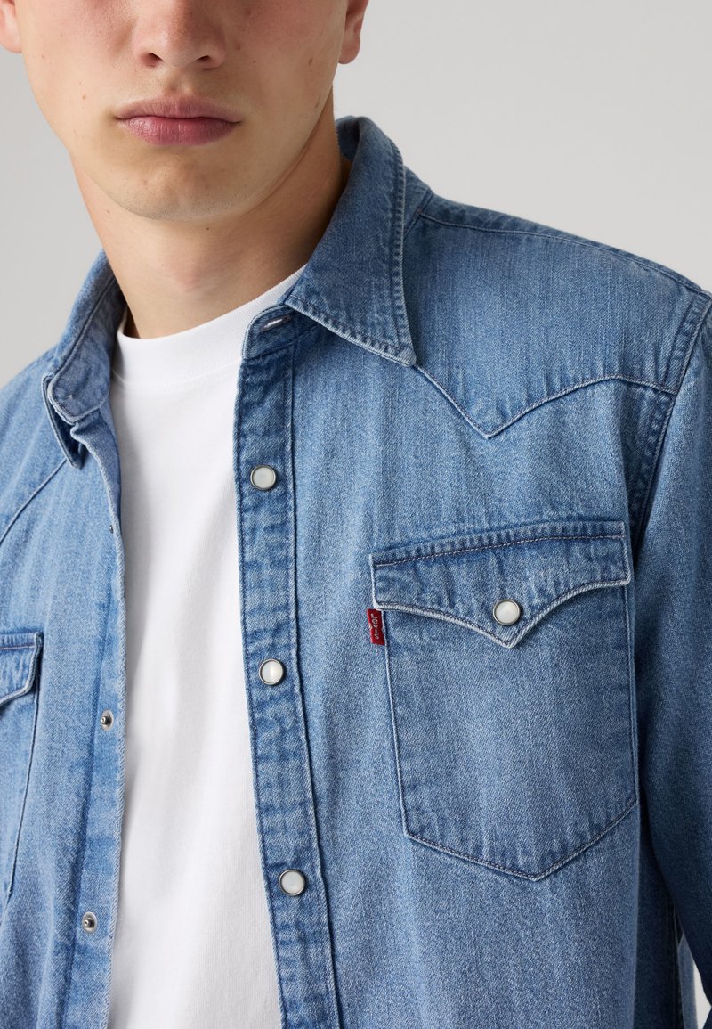 Jeune homme portant une chemise en denim bleu déboutonnée avec des boutons-pression sur un t-shirt blanc devant un fond clair uni.