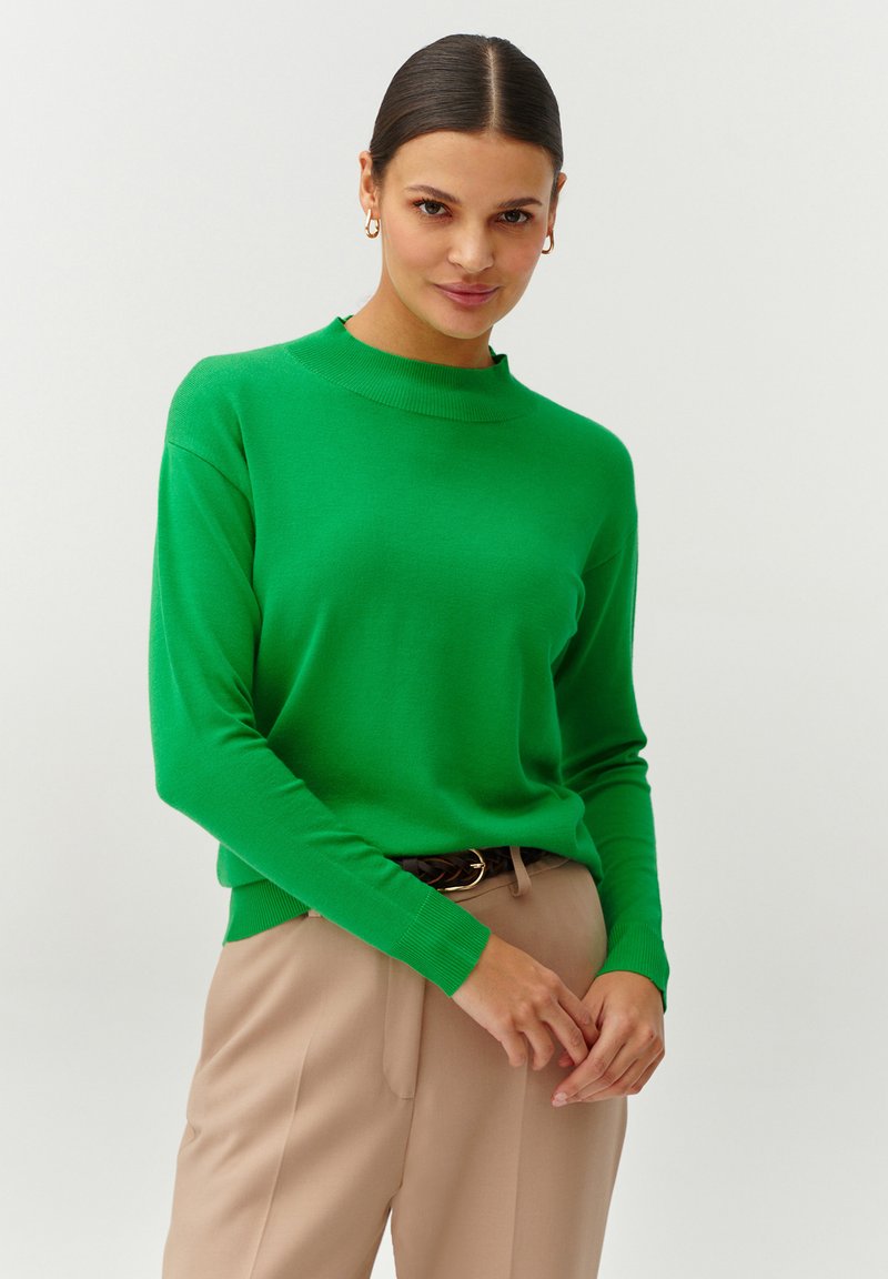 Groene gebreide trui met een hoge halslijn, lange mouwen en geribbelde manchetten, gecombineerd met beige broek. Soepele textuur, lichtgewicht stof.