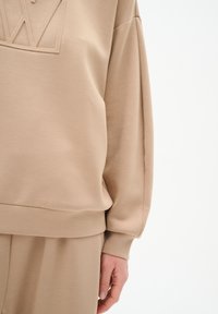 Beige sweatshirt med en texturerad, upphöjd logotypdetalj på bröstet. Har en avslappnad passform, långa ärmar och en mjuk tygtextur.