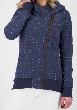 Femme portant un sweat à capuche zippé bleu marine avec petits points blancs, symboles nautiques rouges et un écusson en forme de cœur sur la poitrine, associé à un pantalon blanc.