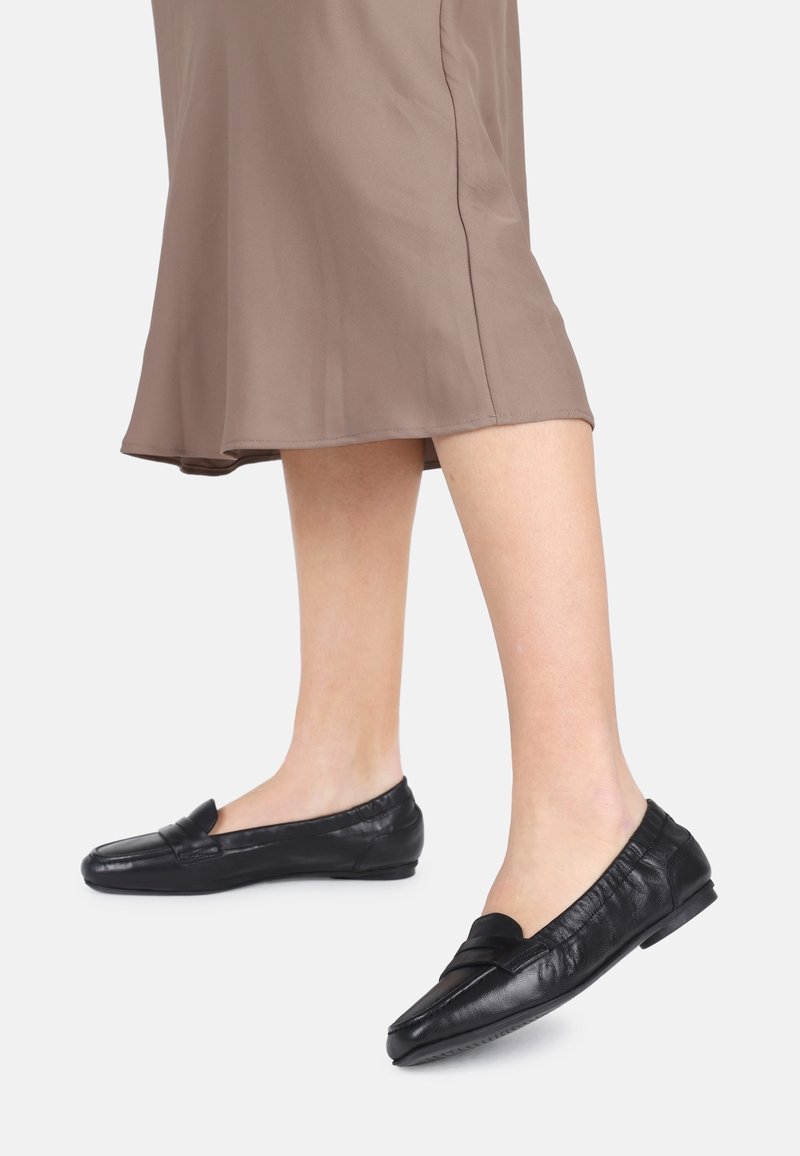 Zwarte leren ballerina's met een strikaccent, laag profiel ontwerp, gladde textuur en minimale stiksels, gecombineerd met een bruine, vloeiende rok.