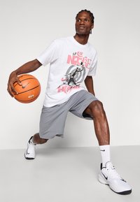 Witte graphic T-shirt met roze en zwarte prints, grijze sportshorts en witte sneakers met zwarte accenten, terwijl hij een basketbal vasthoudt.