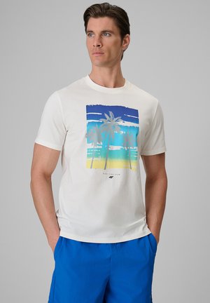 Mężczyzna w białym t-shircie z niebiesko-żółtą grafiką palmy i jasnoniebieskich szortach, stoi z rękami w kieszeniach, patrząc w bok.