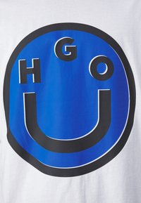 Camiseta blanca con un gráfico circular azul con contornos negros y letras en negrita "HGO" y "U" incorporadas en el diseño.