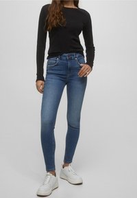 Smalle blauwe jeans met een lichte vervaging, vijf zakken en een knoopsluiting, gecombineerd met een zwart shirt met lange mouwen en witte sneakers.