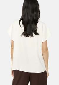 Blusa bianca dalla vestibilità ampia con maniche a sbuffo corte, una texture liscia e un orlo arrotondato. Indossata con pantaloni marroni, vista da dietro.