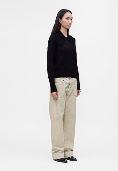 Femme aux longs cheveux foncés portant un haut noir à manches longues avec un col large, un pantalon beige à jambes larges et des chaussures noires pointues, debout de profil.