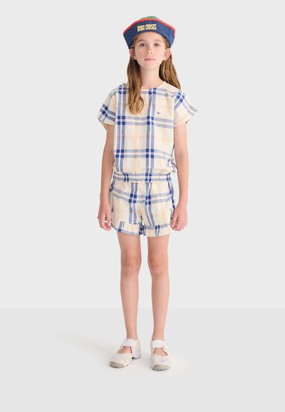 Enfant portant une chemise à manches courtes bleu et beige à carreaux et un short assorti, des chaussures blanches et une casquette colorée avec le texte "BOBO CHOSES".