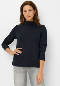 Marineblauwe top met lange mouwen en een hoge hals. Zacht materiaal met een gladde textuur. Voorzien van een gestikte zoom en een comfortabele pasvorm. Draagt lichtgrijze jeans.