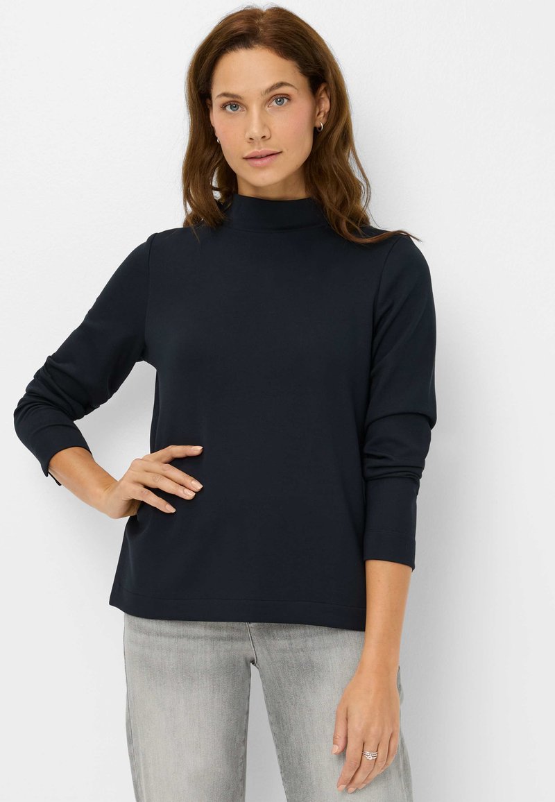 Marineblauwe top met lange mouwen en een hoge hals. Zacht materiaal met een gladde textuur. Voorzien van een gestikte zoom en een comfortabele pasvorm. Draagt lichtgrijze jeans.