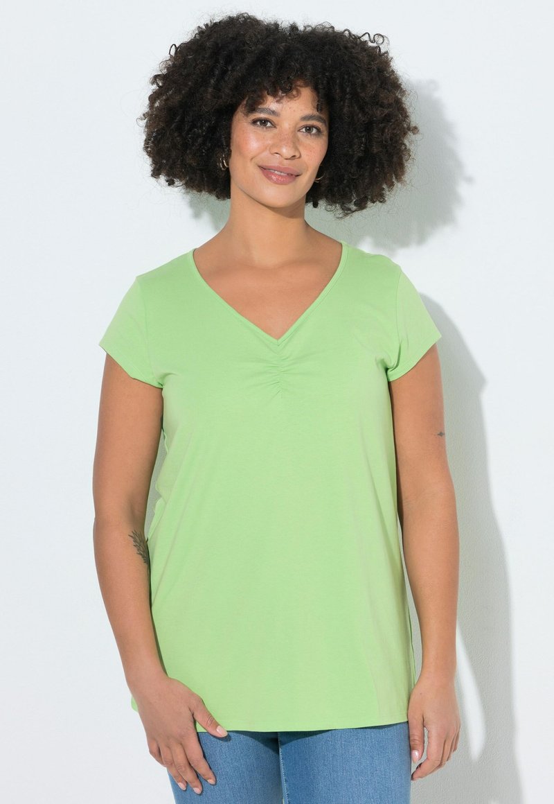 Donna con capelli ricci e scuri, che indossa una maglia a maniche corte color verde chiaro con scollo a V e jeans blu, che sorride leggermente su uno sfondo chiaro.