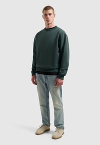 Jonge man staat in een donkergroen sweatshirt, lichtblauwe gescheurde jeans en beige veterschoenen op een effen grijze achtergrond.