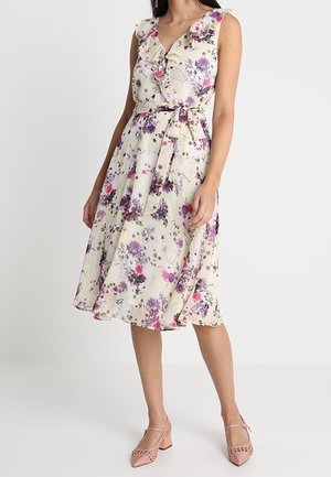 Bezrekawowa kremowa sukienka w fioletowo-różowy kwiatowy wzór, z falbanami przy dekolcie w kształcie litery V, z wiązanym paskiem w talii, noszona z różowymi, teksturowanymi butami na niskim obcasie.