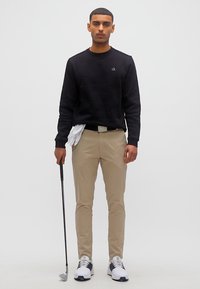 Homme en chemise noire à manches longues et pantalon beige tenant un club de golf dans la main droite, gant blanc glissé dans la ceinture sur un fond blanc uni.