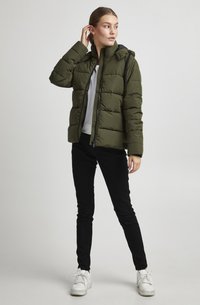 Olivgrön puffjacka med dragkedja fram och huva, med en kviltad design. Tillsammans med svarta byxor och vita sneakers.