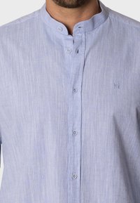 Camisa de botones azul claro en una suave tela texturizada con cuello banda y un pequeño logo bordado en el pecho.