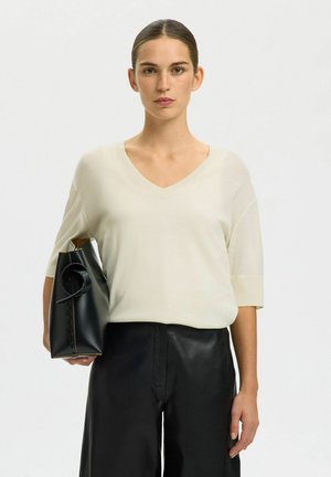 Mujer con el cabello peinado hacia atrás lleva una camiseta crema con escote en V y pantalones negros, sosteniendo un bolso negro de cuero bajo el brazo.