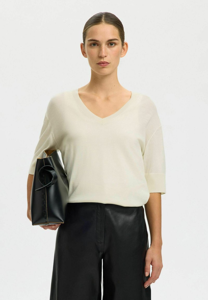 Femme aux cheveux plaqués en arrière portant un haut crème à col en V et un pantalon noir, tenant un sac à main en cuir noir sous le bras.
