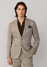 Muž v béžovom obleku s hnedou košeľou a modrým vzorovaným vreckovkým, stojaci s jednou rukou v vrecku pred neutrálnym pozadím.